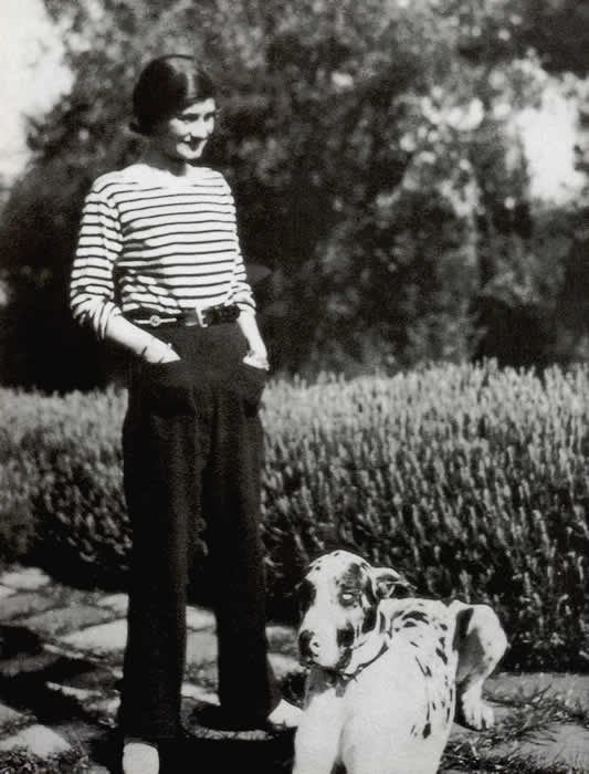 1928年のココ・シャネルの写真。しまのシャツに黒いズボンで、庭に立っている。足もとに大きな犬。撮影者は不明。