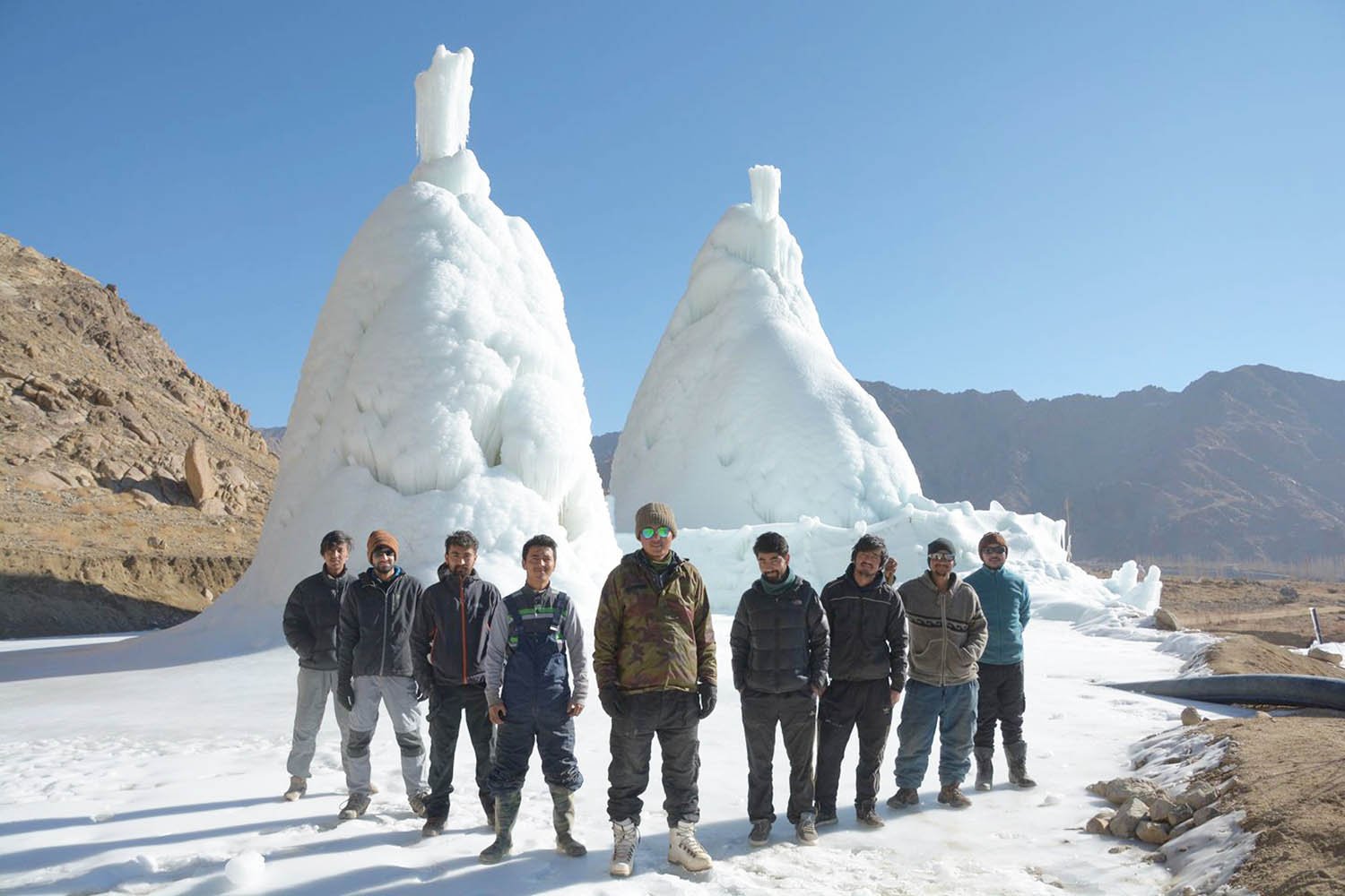乾いた岩山を背に立つ、2基の白い氷の塔。高さは人の背丈の何倍もあり、先からつららのように氷が伸びている。手前に、塔をつくった若者たちが並んでいる。