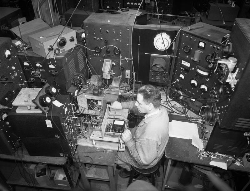 1945年、MITの放射線研究所で、たくさんの計器や機械に囲まれた作業台に向かい、マグネトロンを試験する技術者。白黒写真。