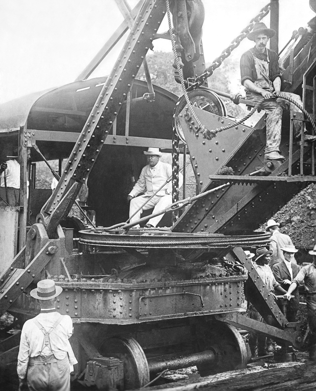 1906年の白黒写真。パナマ運河の工事現場で、巨大な蒸気ショベルの運転席に、白い服と白い帽子のルーズベルト大統領が座っている。まわりに帽子をかぶった作業員たち。